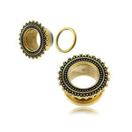A pair of ornate Kaya Movement Flesh Tunnel piercings is shown, resembling cogwheels in design. Predominantly gold with detailed black accents, they bear a striking resemblance to tribal plugs. One stainless steel gauge has its separate parts displayed: the front, the backside, and a small ring that fits around the 19mm opening.