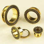 A collection of Kaya Movement's intricately designed Flesh Tunnel piercings, featuring gold and black stainless steel plugs in various sizes, reminiscent of tribal plugs, displayed on a beige background. These ear gauges showcase a beaded pattern and ornamental details around their edges. One of the 19mm gold gauges is shown disassembled.