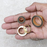 A hand holding three intricate golden rings. Two rings feature a circular design with small black beads outlining the edges, while the third showcases a toothed or gear-like appearance reminiscent of tribal plug designs. These pieces are part of Kaya Movement's collection and include a Flesh Tunnel piercing, Ear gauges, Stainless steel plug, Gold gauges in sizes such as 19mm plugs and 2g gauges. The background is a neutral, textured surface.