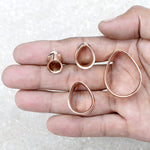 A hand is holding four rose gold metallic shapes resembling components of handcrafted tribal plugs by Kaya Movement: a small strip, two medium-sized oval rings, and one large teardrop ring. They are all placed on the palm against a light-colored background.