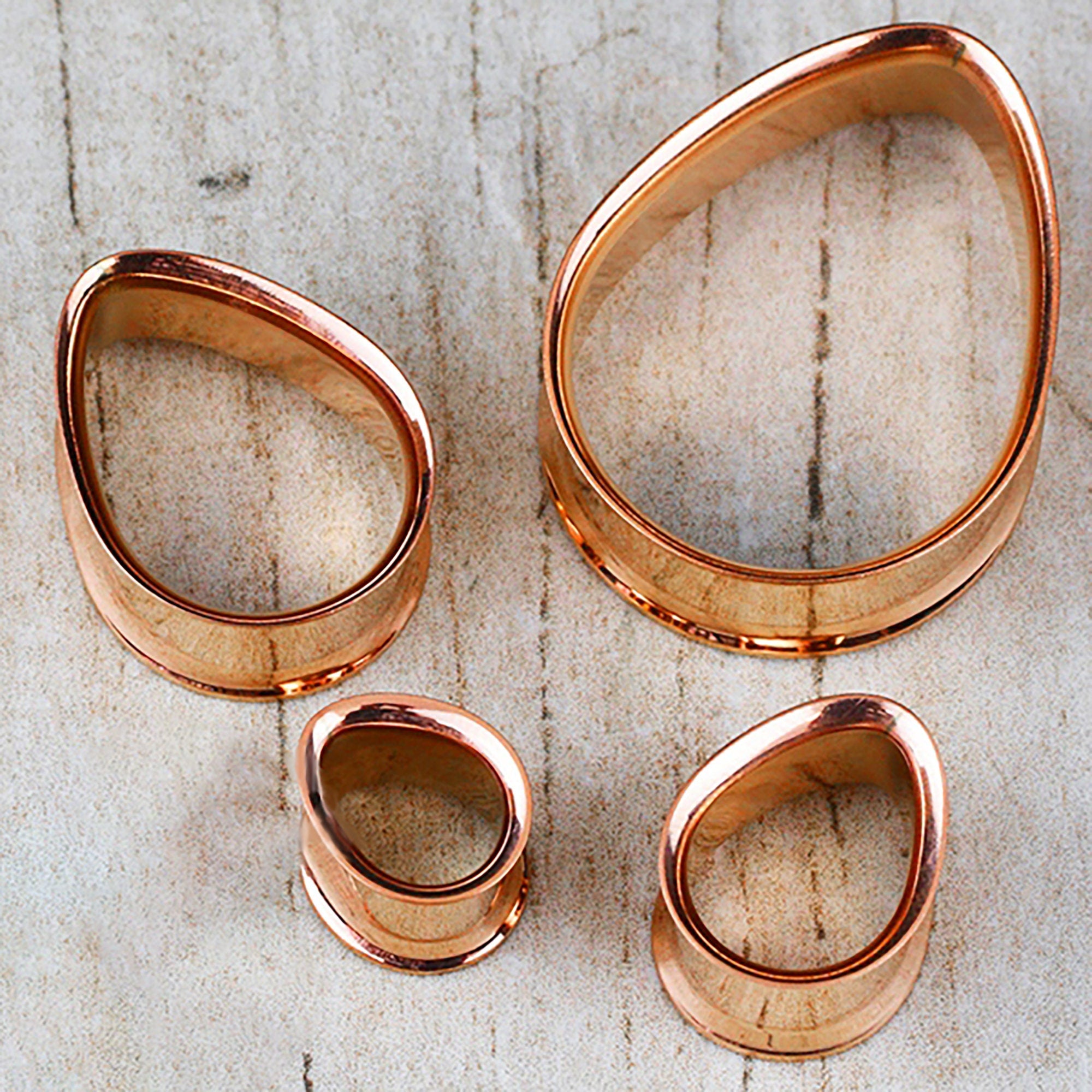 Displayed on a wooden surface, this set of four irregularly shaped metal rings from Kaya Movement—a collection of Teardrop Tunnel Plugs in rose gold—boasts a handcrafted tribal plug design. The 14 gauge teardrop-shaped rings feature smooth edges and a glossy finish that reflects light beautifully.