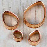 Displayed on a wooden surface, this set of four irregularly shaped metal rings from Kaya Movement—a collection of Teardrop Tunnel Plugs in rose gold—boasts a handcrafted tribal plug design. The 14 gauge teardrop-shaped rings feature smooth edges and a glossy finish that reflects light beautifully.