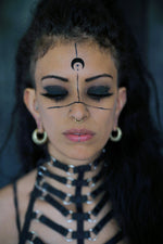 A person with long dark hair and heavy black eyeliner has geometric designs and a moon symbol painted on their face. They have a Kaya Movement Biomechanical 18k Gold Septum Ring in 10mm, large hoop earrings, and are wearing a black strappy outfit. Their eyes are closed, and they appear deep in thought.