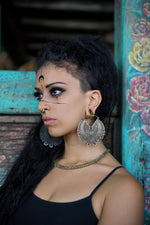 A woman with intricate face paint and large, ornate earrings gazes to the side. She has long, wavy dark hair, which is shaved on one side. Sporting a black top and a gold necklace, she stands against a rustic turquoise wall adorned with floral decorations, her look accentuated by the Kaya Movement's Futuristic Xenomorph 18k Gold-Plated Septum Ring for Men and Women.