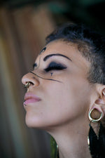 A contemplative individual with a sidecut hairstyle and multiple facial piercings, including the Kaya Movement's Futuristic Xenomorph 18k Gold-Plated Septum Ring for Men and Women, tilts their head back slightly. The septum ring features a biomechanical alien horseshoe design. They have bold eye makeup and intricate black facial tattoos, complemented by large hoop earrings.