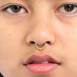A close-up of a person's face focusing on their nose and lips, showcasing the Hinged Teardrop Tribal Septum Ring Clicker by Kaya Movement. The gold septum ring features intricate tribal detailing. The person's light brown skin tone, slightly parted lips, and visible eyes in the background create an exquisite example of stunning nose piercing artistry.