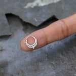 A small, ornate Silver Septum Ring Clicker Hinged Teardrop 16g from Kaya Movement, featuring intricate detailing with tiny beads at the bottom, is balanced on the fingertip of a hand. The background appears to be a weathered, dark surface, highlighting the stunning craftsmanship of this versatile piece suitable for Conch, Daith, Helix Body Piercing or as an 8mm Cartilage Earring.