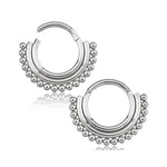 A silver septum clicker, crafted from surgical steel by Kaya Movement, is adorned with small, rounded beads along the outer edge, giving it a decorative and ornate appearance. The hinged design ensures easy wearing. The reflection of the jewelry is visible below it.