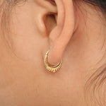 Close-up of a person's ear adorned with a small, intricate gold septum ring clicker from Kaya Movement. The Hinged Teardrop Tribal Septum Ring Clicker 16g features a series of tiny, detailed embellishments along the bottom edge, adding an elegant touch reminiscent of tribal jewelry. Strands of hair are visible near the ear.