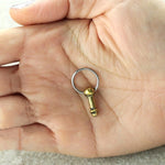 A hand with a subtle septum piercing is holding a small keyring adorned with the "Anatomy Dick Earrings Jewelry" by Kaya Movement, which features a metal pendant shaped like a microphone. The hand is open with the palm facing up, and the keyring lies in the center, gleaming silver while the microphone-shaped pendant glows gold.