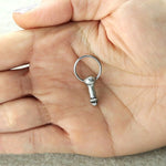 A hand is holding a small keychain resembling the Anatomy Dick Earrings Jewelry Silver from Kaya Movement, crafted from stainless steel 316L. The keychain has a round ring attached for keys, with a light-colored and smooth background surface.