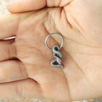 A close-up image of a hand holding a small keychain shaped like an intricately detailed, metallic coiled snake, reminiscent of the exquisite design of Kaya Movement's Silver Snake Hoop Earrings. The keychain loop is attached to the snake's head, echoing the fine craftsmanship seen in septum and helix piercings.