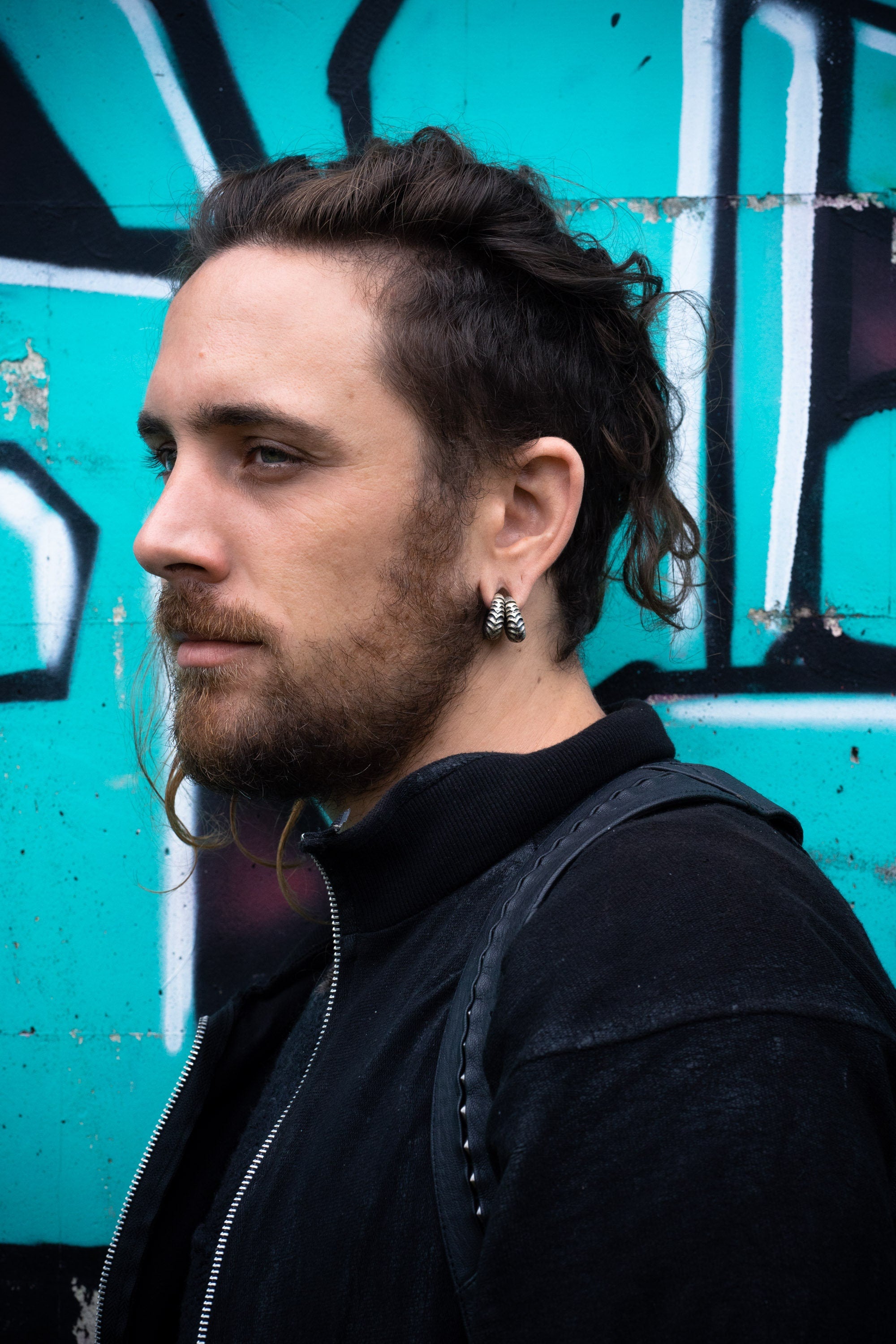A person with a beard and long hair tied back, wearing a black jacket and Kaya Movement's Fractal Alien Bio-Organic Circle Ear Weights in Modern Brass 4mm (6ga) Gauges for stretched lobes, stands in front of a turquoise graffiti wall. The graffiti features black and white abstract shapes.