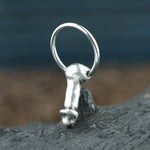A small, silver-colored "Anatomy Dick" pendant from Kaya Movement hangs from a circular ring. The stainless steel 316L pendant is placed on a textured surface with a blurred background.