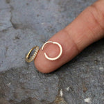 A person holds a small Kaya Movement Hinged Septum Ring Clicker (16g) adorned with tiny cubic zirconia stones, shaped like a hoop, with their index finger. Another identical earring lies on a textured gray surface. These versatile earrings are perfect for nose piercings and various other body piercings such as conch, daith, helix, cartilage, and rook.