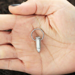 A hand holds one of Kaya Movement's Silver Bullet Gun Dangle Earrings with Charm. The small, silver earring is shaped like a bullet, featuring a loop at the top for attachment. The palm lines and nails of the hand are clearly visible, reminiscent of handling fine septum piercing jewelry in stainless steel. The background is dark and out of focus.