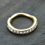 A small gold Hinged Septum Ring Clicker by Kaya Movement, adorned with sparkling cubic zirconia gemstones across the front, is placed on a dark, textured surface. Its design is simple yet elegant, ideal for nose piercings.