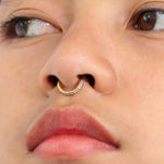 Close-up of a person's face showcasing the Kaya Movement Hinged Septum Ring Clicker 16g. The gold septum ring is adorned with cubic zirconia on one side, enhancing its elegance. The person has light skin, slightly parted lips, and eyes that gaze to the side.
