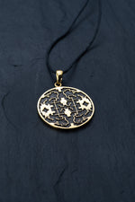 The Shipibo pendant from Kaya Movement, crafted from yellow brass, showcases intricate, maze-like designs and oxidized details. This striking gold pendant hangs elegantly from a black cord against a dark, textured background, featuring symmetrical patterns and geometric shapes that create an eye-catching visual effect.