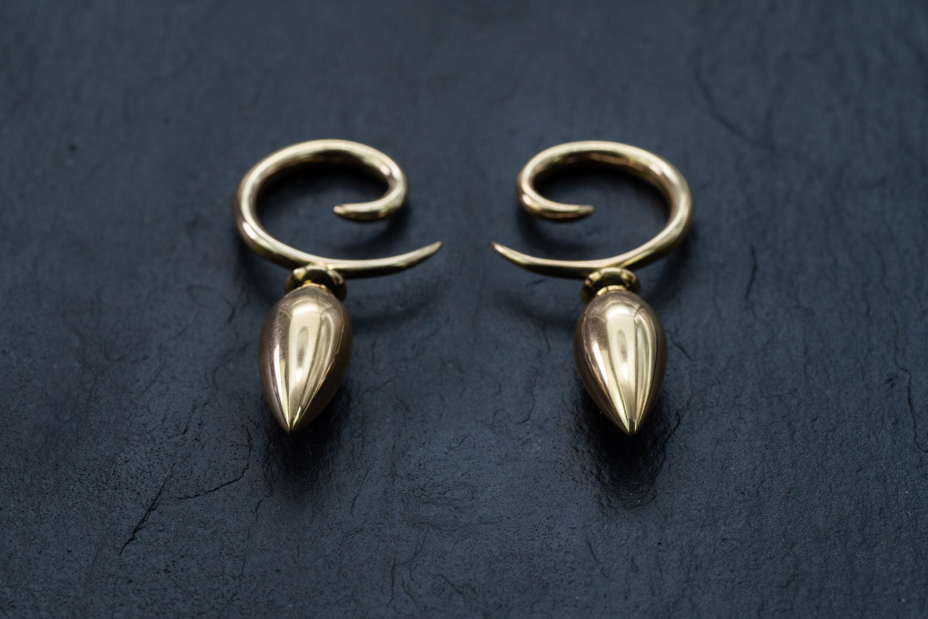 A pair of Kaya Movement Drop Ear Hangers - Spiral Gauges - Ear Weights rests on a dark, textured surface. The gold 4g earrings are identical, showcasing a smooth, polished finish and elegant teardrop-shaped curves designed for stretched ears.
