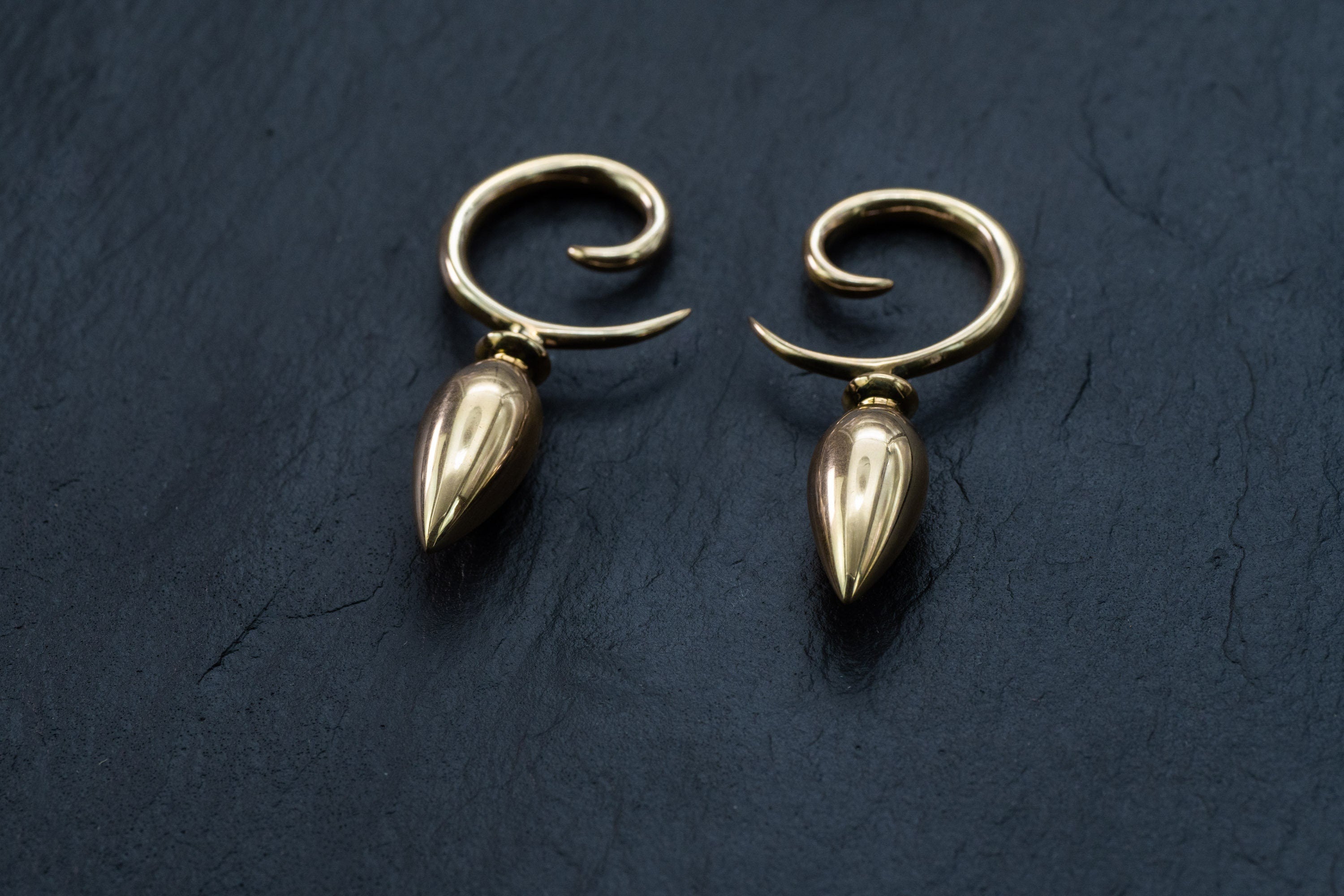 A pair of Kaya Movement's Drop Ear Hangers - Spiral Gauges, featuring a unique hand-carved design in gold, elegantly curve into a hook shape with a sharp downward-pointing tip. Placed on a dark, textured surface, these 4g ear weights for stretched ears highlight their shiny and polished finish.