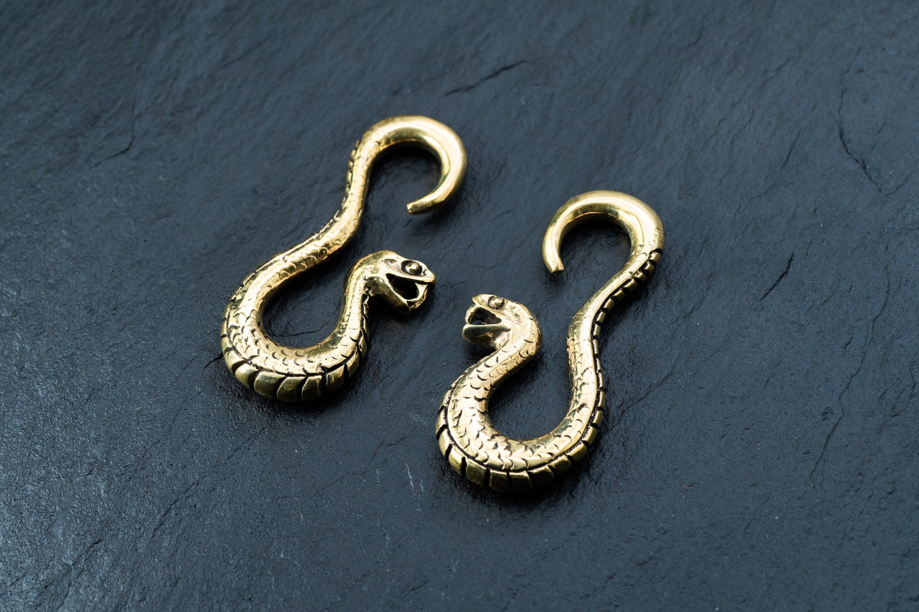 Two Rattlesnake Gold Ear Weights from Kaya Movement rest on a dark slate surface. The ear weights are intricately designed with lifelike scales and open mouths, highlighting the elegance and detailed craftsmanship of these snake brass ear hangers for stretched ears, which resemble antique jewelry.