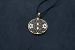 A Shipibo pendant from Kaya Movement, featuring intricate maze-like patterns and oxidized details, hangs from a black cord necklace on a dark, textured surface. The round, yellow brass pendant closely resembles Shipibo Icaros symbols, creating a sophisticated and detailed appearance with geometric shapes and lines.
