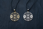 Two circular Shipibo pendants from Kaya Movement, featuring intricate geometric patterns, hang on black cords against a dark background. The left pendant, crafted from yellow brass, showcases oxidized details, while the right one has a silver-toned finish. The symmetrical designs on both shamanic pendants display lines and shapes that contribute to their complex and ornate look.