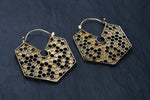 A pair of Kaya Movement's oversized hoop honeycomb hexagon bee gold brass earrings lies on a dark slate surface. These Sacred Geometry Cool Boho Hypercube Jewelry pieces feature a hollowed-out center and showcase an intricate and detailed design in a closed hoop style.