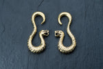 A pair of Kaya Movement's Rattlesnake Gold Ear Weights, intricately designed with detailed scales and open-mouthed heads, are displayed side by side on a dark textured background. These snake-shaped brass ear weights curve elegantly, resembling the shape of question marks.