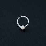 An opal nose ring by Kaya Movement rests delicately on a dark, textured surface. Showcasing septum elegance, this silver 8mm horseshoe design features a seamless and open-ended construction with an 18g small, round opal that adds a subtle touch of iridescent color.