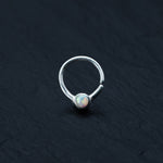 An Opal nose ring from Kaya Movement, specifically the Silver septum ring designed in an 8mm horseshoe seamless style, rests on a dark, textured surface. This refined piece of body jewelry showcases a circular opal gemstone at its center, blending pastel colors to create a subtle iridescent effect that epitomizes septum elegance.