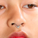 Close-up of a person with light brown skin wearing red lipstick, adorned with a Kaya Movement Opal Nose Ring. The elegant silver septum ring is centered with a small iridescent jewel. The person's eyes are partially visible, gazing to the side.