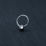 The Zircon Silver septum ring by Kaya Movement, an 8mm small seamless nose ring designed as a horseshoe piercing in 18g, features a minimalist silver design with a clear Crystal Zircon at its center. Resting elegantly on a dark slate surface, this piece of body jewelry embodies subtlety and sophistication.