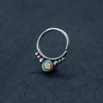 A close-up image of the Kaya Movement Opal Septum Ring Silver, featuring a translucent opal stone at its center in a teardrop design, surrounded by small silver beads. This seamless 18g septum jewelry is positioned on a dark, textured surface, accentuating the intricate details and colorful reflections of the 8mm opal.