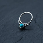 A Kaya Movement silver septum ring with an open band design, adorned with small decorative beads and featuring a central turquoise gemstone, is photographed against a dark, textured background.