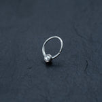 A delicate Zircon Silver septum ring by Kaya Movement, featuring a single clear Crystal Zircon at its center, is positioned on a dark textured surface, highlighting the intricate simplicity of this 18g Horseshoe piercing nose jewelry.