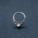 Close-up of the Zircon Silver Nose Septum Ring Piercing by Kaya Movement, showcasing its unique circular design. This sterling silver piece features a prominent central zircon gem, surrounded by small spherical accents on a dark, textured surface. The cute 8mm teardrop shape adds a touch of goth-inspired elegance to this daith jewelry.