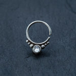 The Zircon Silver Nose Septum Ring Piercing by Kaya Movement, a small, teardrop-shaped septum nose ring with a decorative design, is shown on a dark textured surface. The ring features a clear, round gemstone at the bottom, flanked by a row of small silver beads, adding an intricate and elegant touch.