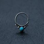 A stunning septum ring from Kaya Movement is showcased against a black textured background, featuring 925 silver and a captivating turquoise gemstone at its center. The intricately designed ring boasts tiny silver beads encircling the vibrant stone, exuding a delicate Bohemian charm that makes it perfect for an ear piercing such as a cartilage, daith, or helix piercing.