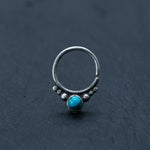 The Kaya Movement's Silver Septum Ring with Turquoise, also suitable as a cartilage earring, daith piercing, or helix piercing, features a turquoise gemstone bead at the center, flanked by small decorative silver dots. Displayed against a dark textured background, the open-ended design ensures it can be easily worn. This piece makes for an elegant Bohemian jewelry gift.