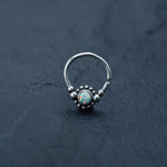 A Kaya Movement silver septum ring, featuring a central opal gemstone encircled by small silver beads, is positioned on a dark, textured surface. The iridescent opal showcases a mix of vibrant colors. This piece, suitable for use as a cartilage earring, daith piercing, or helix piercing, also makes an exquisite Bohemian jewelry gift.