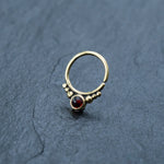 A small, elegant Kaya Movement Garnet Teardrop septum ring featuring a central garnet stone and two clusters of three gold beads on either side, resting on a textured dark grey surface.