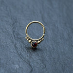 The Kaya Movement Garnet Teardrop Septum features a seamless 8mm gold-colored brass nose ring, elegantly adorned with a small red garnet gemstone and symmetrically arranged gold beads, displayed against a dark gray textured background.