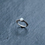 A delicate 925 silver nose ring by Kaya Movement, featuring a round white opal gemstone, is placed on a textured grey surface. The thin band has small decorative ridges, adding to the elegant and understated design of this septum, rook piercing jewelry piece.