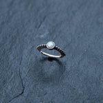 A delicate opal jewelry piece, this 925 silver nose ring by Kaya Movement features a circular opal prominently displayed in an 8mm septum design and is showcased against a dark, textured slate background. The silver band boasts a detailed beaded design, adding an element of intricacy to the overall aesthetic.
