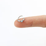 A small, 925 silver nose ring from Kaya Movement, featuring a central Opal bead, is balanced on the tip of an index finger against a plain white background. This 8mm septum ring, ideal for piercings like rook, tragus, and helix jewelry, boasts a textured design around the bead.