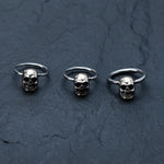 Three Skull Ball Closure Rings, adorned with detailed small skulls as their centerpieces, are neatly arranged in a row on a dark, textured surface. These Stainless Steel 316L rings from Kaya Movement add an edgy touch to your look and are perfect for use as jewelry or piercings, available in 18g, 16g, and 14g sizes.