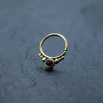 A delicate Garnet Teardrop septum ring by Kaya Movement is displayed on a dark gray textured surface. This 8mm gold septum ring, reminiscent of a small simple brass septum piece, features a deep red garnet gemstone at its center, adorned by six tiny gold beads symmetrically arranged on either side.