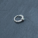 Close-up image of the Kaya Movement 925 silver nose ring with Opal - 8mm septum, lying on a dark, textured surface. This exquisite silver nose hoop features intricate detailing and a single opalescent stone, capturing the essence of refined piercings for rook, tragus, and helix jewelry.